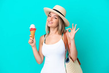 Elinde dondurma ve plaj çantası olan genç Romen kadın mavi arka planda izole edilmiş ve parmaklarıyla tamam işareti gösteriyor