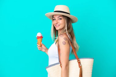 Elinde dondurma ve plaj çantası olan genç Romen kadın mavi arka planda izole edilmiş gülümsüyor.
