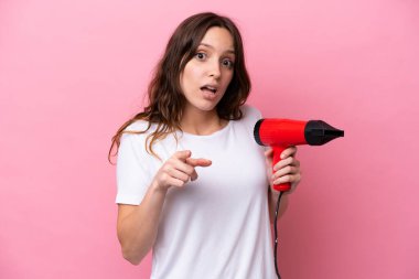 Pembe arka planda saç kurutma makinesi tutan beyaz bir kadın şaşırmış ve ön tarafı işaret ediyor.