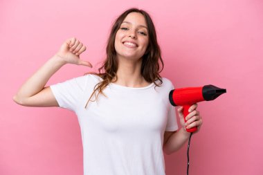 Pembe arka planda saç kurutma makinesi tutan beyaz bir kadın gururlu ve kendini beğenmiş.
