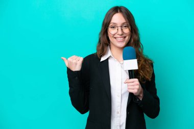Young TV presenter woman isolated on blue background pointing to the side to present a product