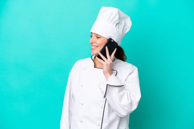 Kafkasyalı genç aşçı kadın mavi arka planda izole edilmiş cep telefonuyla biriyle sohbet ediyor.
