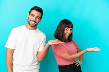 Mavi arka planda izole edilmiş genç bir çift geriye dönüp bir ürün sunuyor.