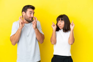Sarı arka planda izole edilmiş genç bir çift parmaklarını çapraz yapıp en iyisini diliyor.