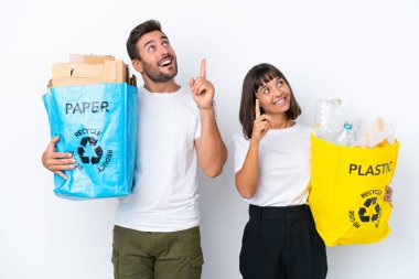 Beyaz arka planda izole edilmiş plastik ve kağıt dolu bir çanta taşıyan genç çift işaret parmağıyla işaret ediyor. Harika bir fikir.