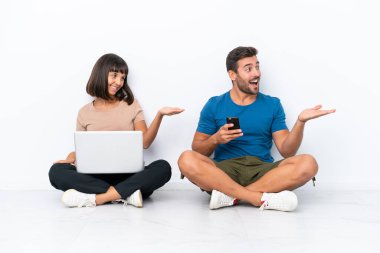 Yerde oturan genç çift beyaz arka planda izole edilmiş bilgisayar ve cep telefonlarını tutarken geriye dönüp bir ürün sunuyorlar.