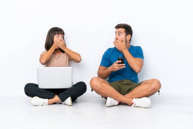 Yerde oturan genç çift, ellerinde cep telefonu ve cep telefonuyla beyaz arka planda izole edilmiş ağızlarını uygunsuz bir şey söyledikleri için elleriyle kapatıyor.