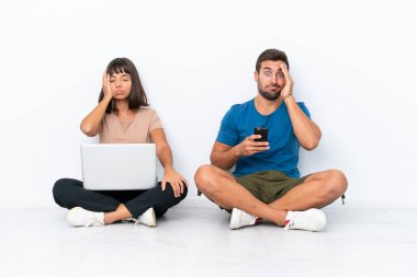 Yerde oturan genç bir çift ellerinde bilgisayar ve cep telefonuyla beyaz arka planda şaşırmış ve şok olmuş bir yüz ifadesiyle izole edilmiş.