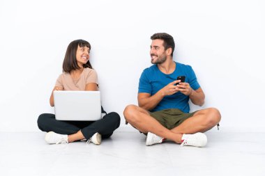 Young couple sitting on the floor holding pc and mobile phone isolated on white background looking over the shoulder with a smile