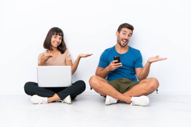Young couple sitting on the floor holding pc and mobile phone isolated on white background extending hands to the side for inviting to come