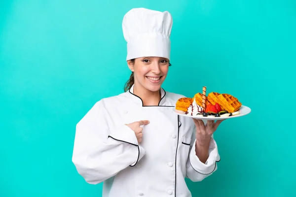 Young caucasian chef woman holding waffles isolated on blue background with surprise facial expression