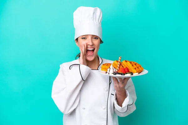 Young caucasian chef woman holding waffles isolated on blue background shouting with mouth wide open
