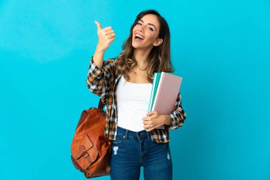 Genç bayan öğrenci, mavi arka planda izole edildi. Başparmakları havada, çünkü iyi bir şey oldu.