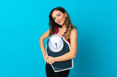 Mavi arka planda kolları kalçasında ve tartı makinesini tutan beyaz bir kadın.