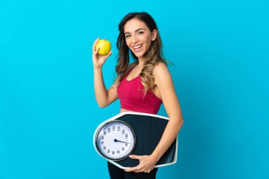 Mavi arka planda tartı tutan beyaz bir kadın. Tartı makinesiyle ve elmayla izole edilmiş.