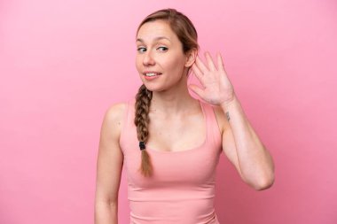 Pembe arka planda izole edilmiş genç beyaz kadın elini kulağına koyarak bir şeyler dinliyor.