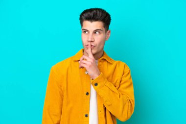 Genç, beyaz, mavi bakkalda izole edilmiş genç bir adam sessizlik işareti gösteriyor. Parmağını ağzına sok.