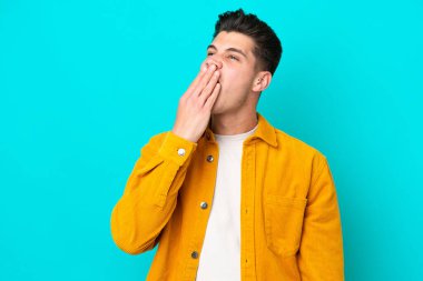 Genç, yakışıklı, beyaz bir adam, mavi bir fırında izole edilmiş, esniyor ve ağzını eliyle kapatıyor.