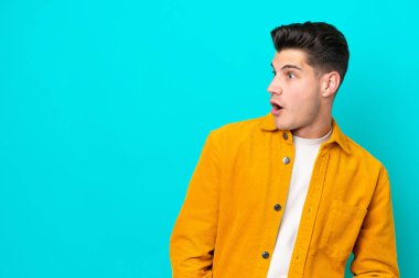 Genç, yakışıklı, beyaz bir adam, mavi bakkalda izole edilmiş. Yan tarafa bakarken sürpriz jest yapıyor.