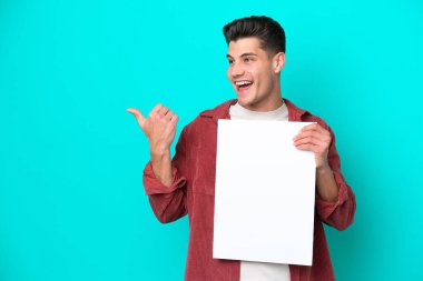 Genç, yakışıklı, beyaz bir adam mavi bir bakkalda izole edilmiş. Elinde boş bir levha var ve onu işaret ediyor.