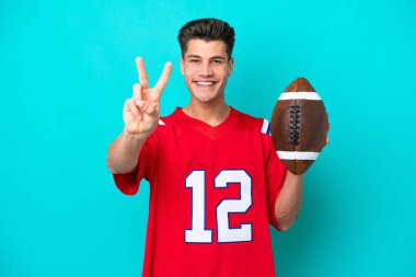 Genç beyaz adam rugby oynuyor. Mavi arka planda izole edilmiş. Gülümsüyor ve zafer işareti gösteriyor.