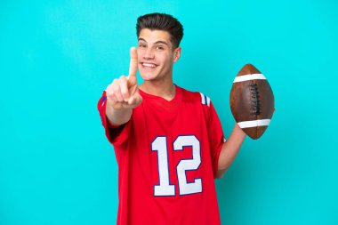 Genç beyaz adam rugby oynuyor. Mavi arka planda izole edilmiş. Parmağını kaldırıyor ve gösteriyor.