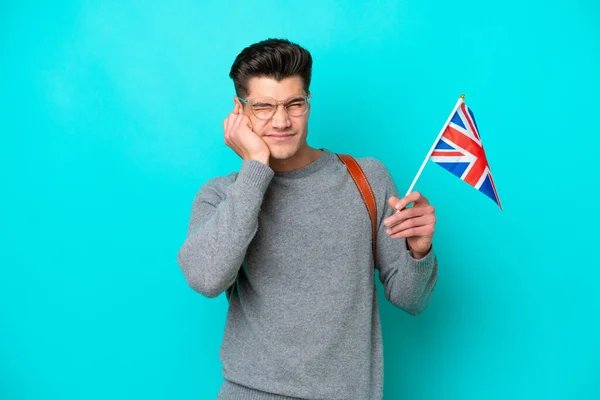 Young caucasian man holding an United Kingdom flag isolated on blue background frustrated and covering ears