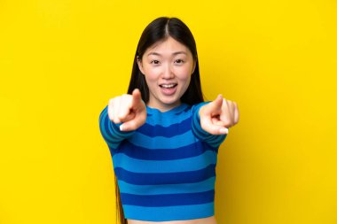 Sarı arka planda izole edilmiş genç Çinli kadın şaşırmış ve ön tarafı işaret ediyor.