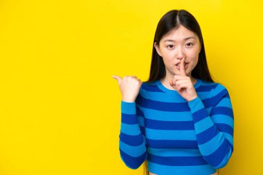 Young Chinese woman isolated on yellow background pointing to the side and doing silence gesture