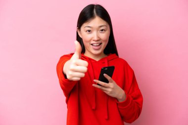 Pembe arka planda izole edilmiş genç Çinli bir kadın başparmak kaldırırken cep telefonu kullanıyor.