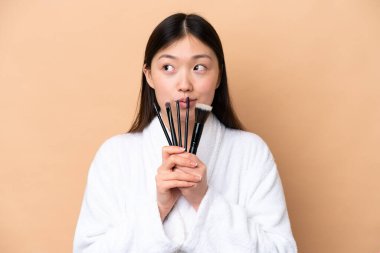 Genç Çinli bir kadın bej arka planda izole edilmiş elinde makyaj fırçası ve yukarı bakıyor.