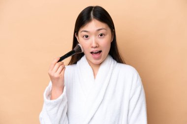 Genç Çinli kadın bej arka planda izole edilmiş elinde makyaj fırçası ve şaşırmış