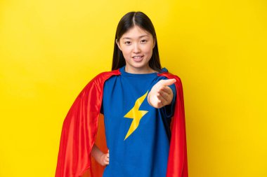 Young Chinese woman isolated on yellow background in superhero costume and making a deal