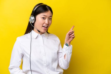 Tele pazarlamacı Çinli kadın sarı arka planda izole edilmiş bir kulaklıkla çalışıyor. Parmakla göstererek bir ürün sunuyor.