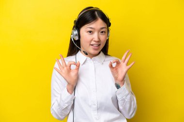 Tele pazarlamacı Çinli kadın sarı arka planda izole edilmiş kulaklıkla çalışıyor. Parmaklarıyla 