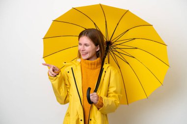 Yağmura dayanıklı ceketi ve şemsiyesi olan genç bir İngiliz kadın beyaz arka planda izole edilmiş bir şekilde parmak ucunda bir ürün sunuyor.