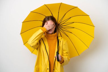 Young English woman with rainproof coat and umbrella isolated on white background covering eyes by hands. Do not want to see something
