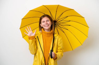 Yağmura dayanıklı ceketi ve şemsiyesi olan genç İngiliz kadın beyaz arka planda beş parmaklı sayım yapıyor.