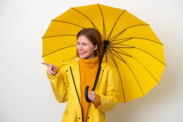 サイドに指を指し 製品を提示白い背景に隔離レインプルーフコートと傘を持つ若い英語の女性 — ストック写真