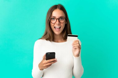 Genç bir kadın cep telefonuyla mavi arka plan alışverişi yapıyor ve elinde şaşırmış bir ifadeyle kredi kartı tutuyor.
