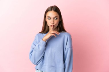 Soyutlanmış pembe arka planda genç bir kadın sessizlik işareti gösteriyor. Parmağını ağzına sok.