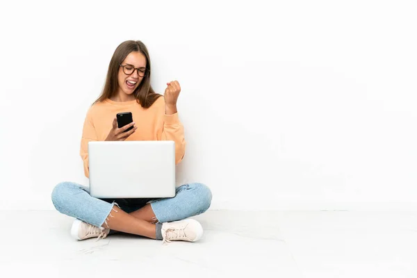 Dizüstü Bilgisayarı Olan Genç Bir Kadın Yerde Telefonuyla Zafer Pozisyonunda — Stok fotoğraf