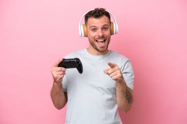 Young Brazilian man playing with video game controller isolated on pink background surprised and pointing front