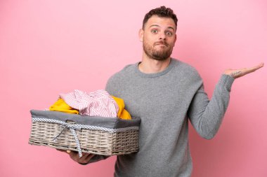 Genç Brezilyalı bir adam pembe arka planda izole edilmiş bir çamaşır sepetini tutarken ellerini kaldırırken şüphe duyuyor.