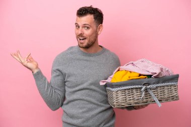 Genç Brezilyalı bir adam pembe arka planda izole edilmiş bir çamaşır sepeti tutuyor. Davet etmek için ellerini yana uzatıyor.