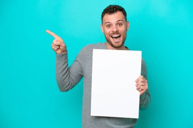 Young Brazilian man holding an empty placard isolated on blue background holding an empty placard and pointing side