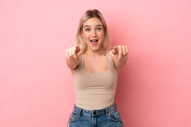 Pembe arka planda izole edilmiş genç beyaz kadın şaşırmış ve ön tarafı işaret ediyor.