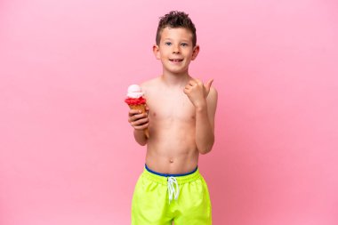 Pembe arka planda izole edilmiş dondurma yiyen beyaz bir çocuk bir ürün sunmak için yan tarafı işaret ediyor.