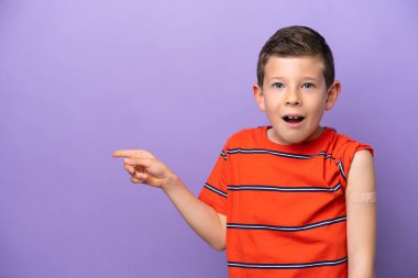 Little boy wearing a band aid isolated on purple background surprised and pointing finger to the side