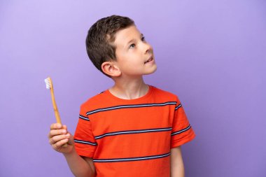 Little boy brushing teeth isolated on purple background looking up while smiling